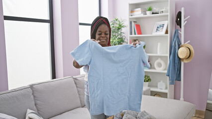 Cheerful african american woman beaming with confidence, holding t-shirt, enjoying her time indoors...