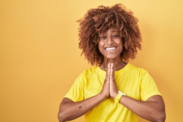 Young hispanic woman with curly hair standing over yellow background praying with hands together...