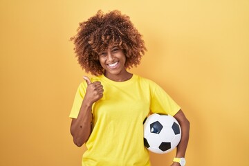 Young hispanic woman with curly hair holding football ball smiling happy and positive, thumb up doing excellent and approval sign