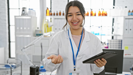 A smiling young woman in a lab coat holding a tablet stands in a pharmacy with shelves of medication in the background.