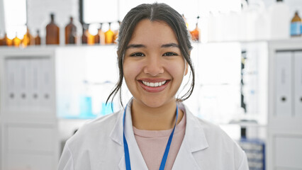 A smiling young hispanic woman in a white lab coat standing in a pharmacy.