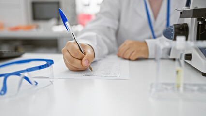Professional woman in lab coat writing on a form, with microscope and eyeglasses in a clinical...