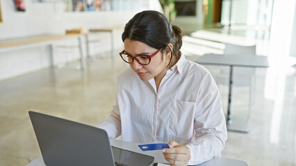 A focused hispanic woman in an office holding a credit card while working on a laptop, embodying professionalism and finance.