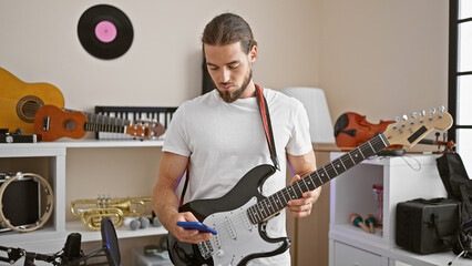 Young hispanic man musician playing electrical guitar using smartphone at music studio