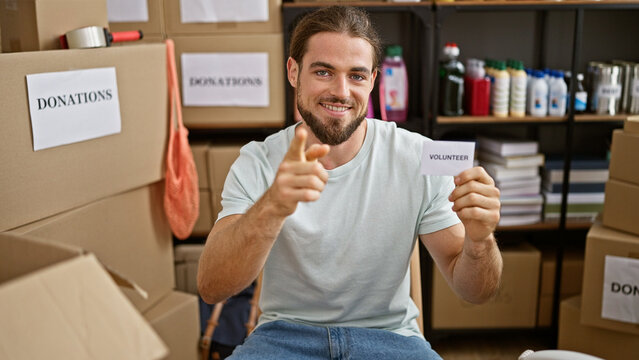 Young hispanic man holding volunteer paper pointing to camera at charity center