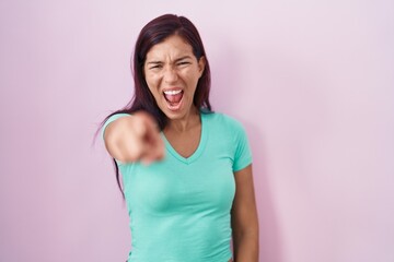 Young hispanic woman standing over pink background pointing displeased and frustrated to the...