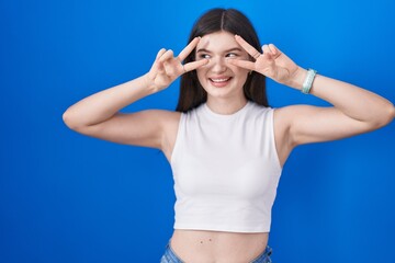 Fototapeta na wymiar Young caucasian woman standing over blue background doing peace symbol with fingers over face, smiling cheerful showing victory