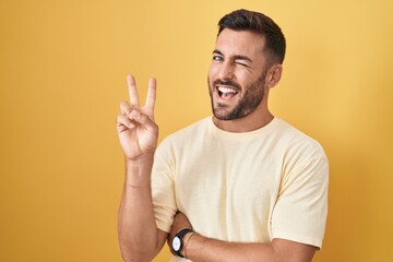 Handsome hispanic man standing over yellow background smiling with happy face winking at the camera...