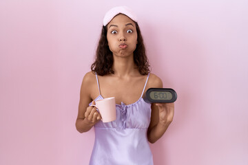 Young hispanic woman wearing nightgown holding alarm clock puffing cheeks with funny face. mouth...