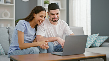 Cheerful, beautiful couple in love sitting together on a cozy sofa at home, smiling while using their laptop in their comfortable living room setting