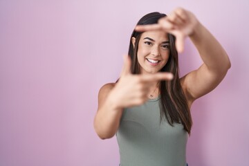 Fototapeta premium Hispanic woman standing over pink background smiling making frame with hands and fingers with happy face. creativity and photography concept.
