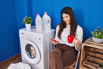 Young beautiful hispanic woman using smartphone drinking coffee waiting for washing machine at laundry room