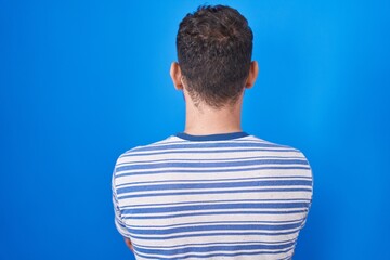 Young hispanic man standing over blue background standing backwards looking away with crossed arms