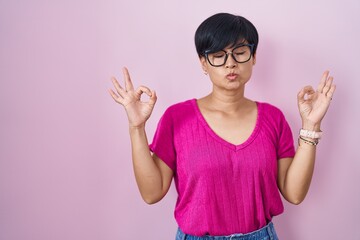 Young asian woman with short hair standing over pink background relaxed and smiling with eyes closed doing meditation gesture with fingers. yoga concept.