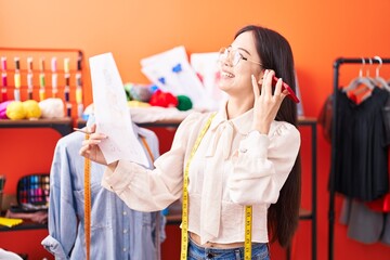 Young chinese woman tailor listening voice message by smartphone looking clothing design at atelier