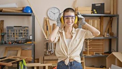 A smiling young woman wearing safety goggles and ear protection in a well-equipped carpentry...