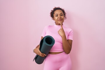 Young hispanic woman with curly hair holding yoga mat over pink background thinking concentrated about doubt with finger on chin and looking up wondering
