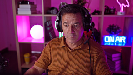 Serious middle-aged man with headphones in a colorful gaming room at night conveys vibrant leisure...