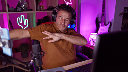 Middle-aged man streaming in a neon-lit gaming room wearing headphones gesturing at his computer monitor.