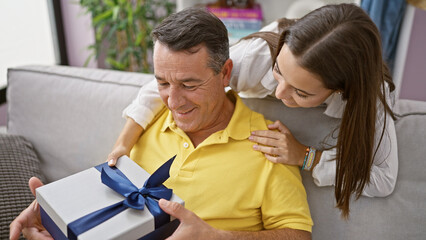 At home, a surprised father and daughter smiling together, sitting on the sofa with a gift in a...
