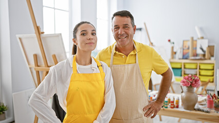 Two smiling artists, a man and woman, confidently standing together by their canvas at an indoor art studio