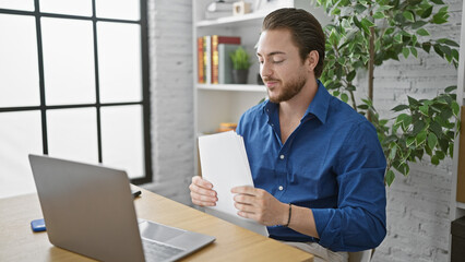 Young hispanic man business worker reading document using laptop at the office