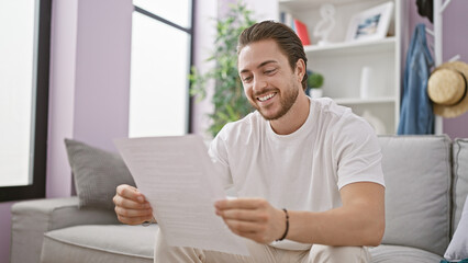 Fototapeta na wymiar Young hispanic man reading document sitting on sofa smiling at home