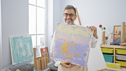 Joyful young hispanic male artist with grey-haired beard, wearing glasses and apron, confidently...
