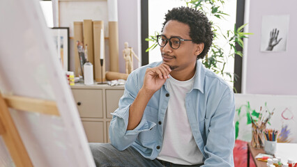 Thoughtful man with glasses in art studio contemplates his painting in a creative indoor setting.