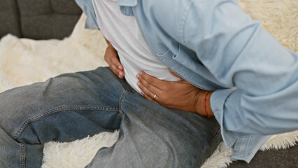 A man experiencing abdominal pain while sitting on a couch indoors, clutching his stomach