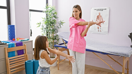 A woman pediatrician demonstrates exercise to a young girl patient in a bright therapy clinic room.
