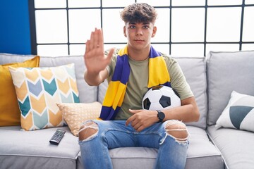 Hispanic teenager sitting on the sofa watching football match with open hand doing stop sign with...