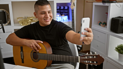 Smiling young latin man, an artist in his prime, striking a melody on his classical guitar, takes a selfie with his smartphone in a music studio