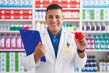 Young hispanic man working at pharmacy drugstore holding heart smiling with a happy and cool smile...