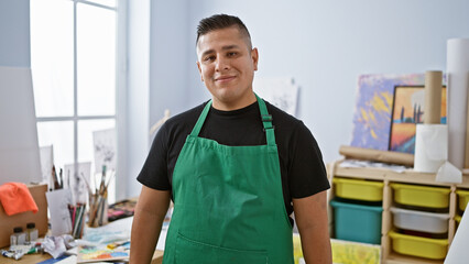 Confident young latin man artist, apron-adorned and brush in hand, standing proudly in his art...