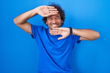 Hispanic young man standing over blue background smiling cheerful playing peek a boo with hands...