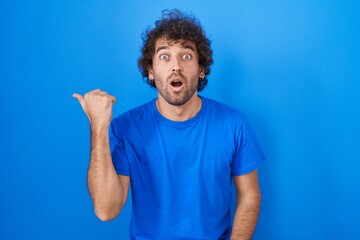 Hispanic young man standing over blue background surprised pointing with hand finger to the side, open mouth amazed expression.