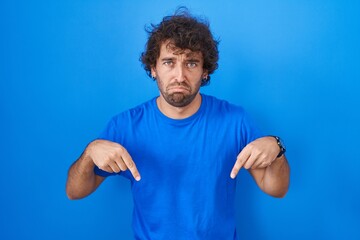 Hispanic young man standing over blue background pointing down looking sad and upset, indicating...