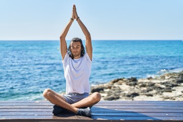 Young hispanic man doing yoga exercise sitting on bench at seaside