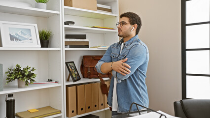 A young man with beard wearing wireless earphones stands thoughtfully in a modern office interior.