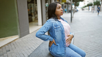 Tired young pregnant woman, touching her belly, sits in contemplation on an urban street bench,...