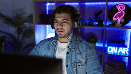 A man with headphones sits in a dark, neon-lit gaming room at night, focused on an unseen screen.