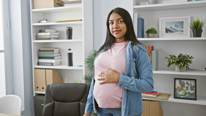 Smiling young pregnant woman confidently touches belly while working in the office, embodying maternity and business success.