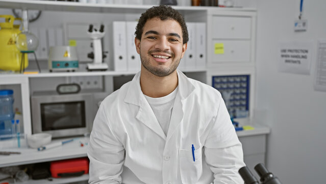 Smiling young man in a white lab coat standing in a laboratory setting with equipment and microscope.