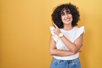 Young middle east woman standing over yellow background with a big smile on face, pointing with hand and finger to the side looking at the camera.