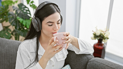 A serene hispanic woman enjoys coffee and music on headphones in a cozy living room setting.