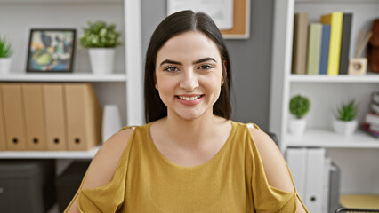 Smiling young woman in casual office attire seated indoors with a neat shelf background.