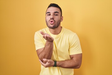 Young hispanic man standing over yellow background looking at the camera blowing a kiss with hand on air being lovely and sexy. love expression.