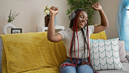 Joyful african american woman, with beautiful braids, dancing on her sofa at home, grooving to the music she's listening to on her headphones. enjoying her own fun-filled indoor lifestyle.