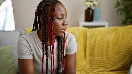 Beautiful african american woman sitting on living room sofa, sporting braids and a serious...